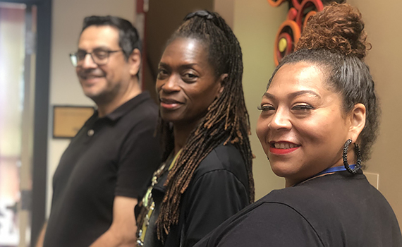 Closeup of three BIPOC coworkers, two women and one man, standing side-by-side with heads turned to smile at the camera.