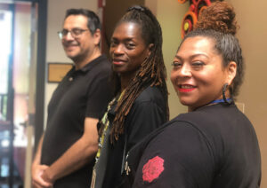 Closeup of three BIPOC coworkers, two women and one man, standing side-by-side with heads turned to smile at the camera.