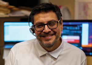 A man with dark hair and glasses talks with a headset smiles into the camera. There are 2 computer monitors behind him.