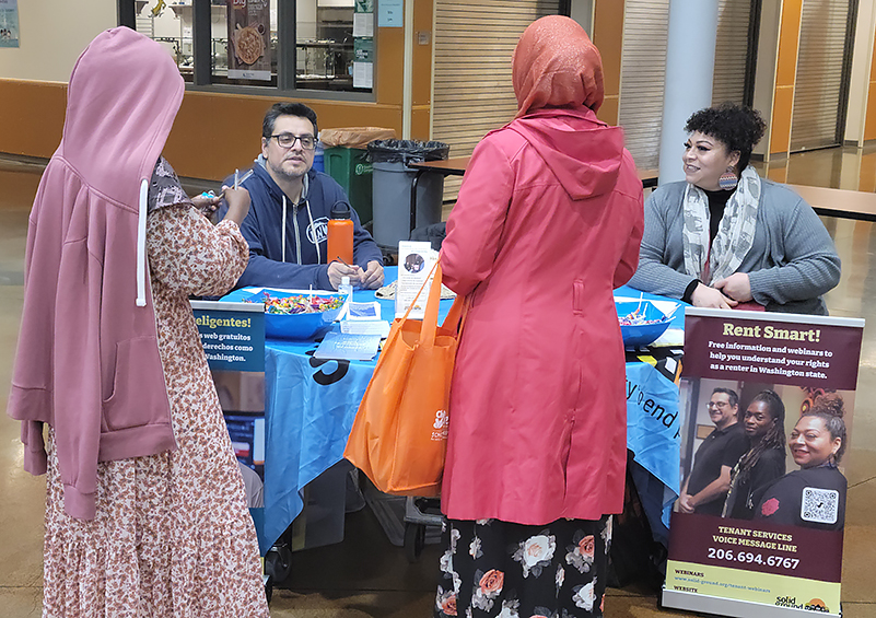 Two women in colorful dresses and headscarves talk to a man and woman seated at table with various outreach materials on it. A banner that reads Rent Smart is next to the table on the right side.