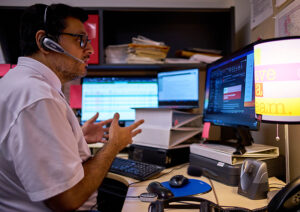 Latino man with dark glasses, hair, and facial hair – wearing a headset and a white shirt – sits at a desk in front of a computer monitor. He’s talking and gesturing with his hands.