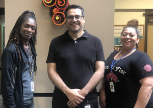 Three BIPOC coworkers, a man and two women, pose in a row, smiling.