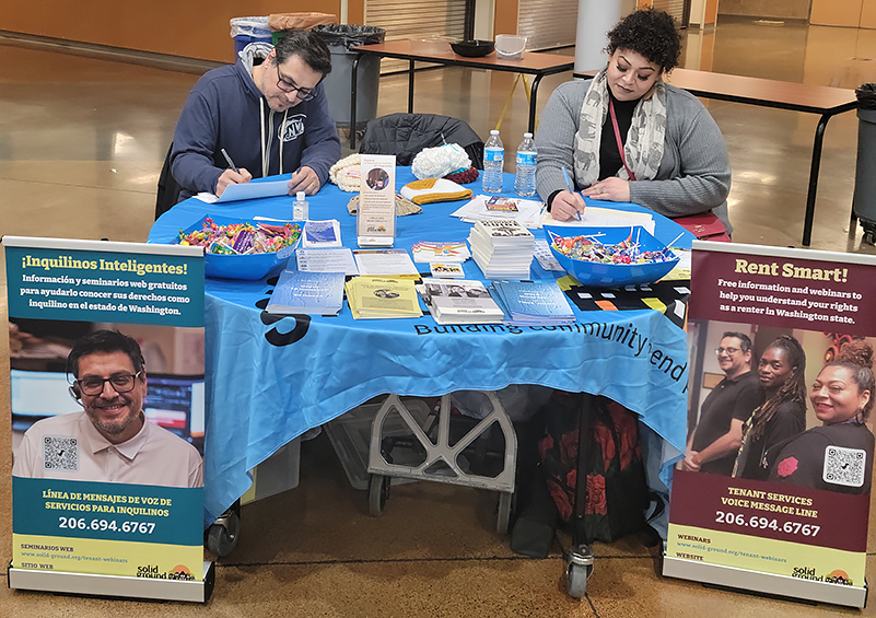 A man and woman are seated at table with various print outreach materials on it. There are pullup banners with Rent Smart information on either side of the table in English (right) and Spanish (left).