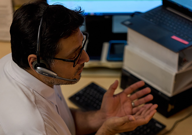 A man with dark hair and glasses talks with a headset on in front of a computer. He gestures with this hands.