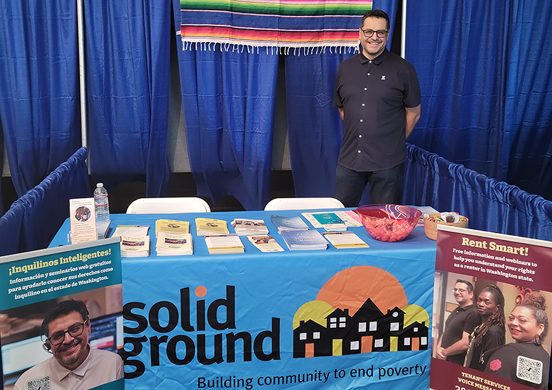 A man wearing all black and glasses stands behind a table with a blue tablecloth with the Solid Ground logo on it. There are print materials on the table, and pullup banners with Rent Smart information on either side in English (right) and Spanish (left).