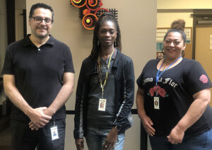 Three BIPOC coworkers, a man and two women, pose in a row, smiling.