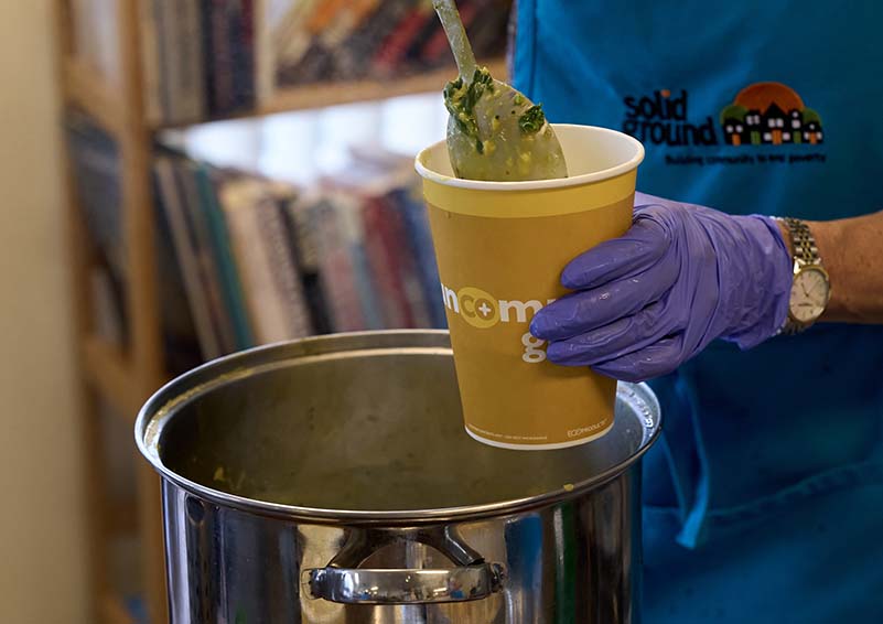 A close-up shot of someone in a blue Solid Ground apron spooning steaming split-pea soup from a pot into a to-go cup.