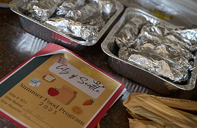 Two alumnium trays full of foods also wrapped in aluminum foil next to a sign that reads, "City of Seattle, Summer Food Program, 2023."