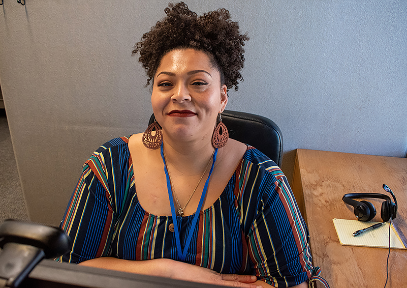 Black woman with curly hair piled on top of her head and wearing a colorful striped top sits in a cubicle in front of two computer screens. She smiles into the camera. There's a headset, yellow note pad, and pen on the desk to her left.