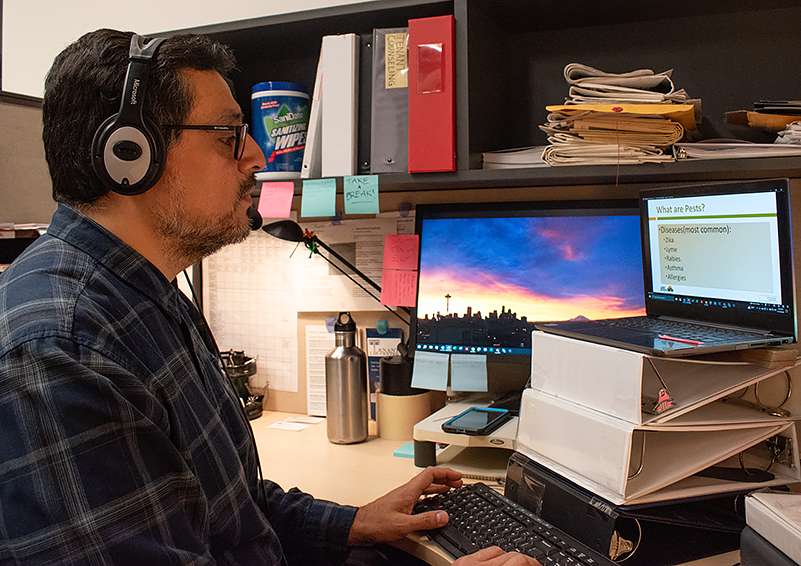 Latino man with dark glasses, hair, and facial hair – wearing a headset and a blue plaid shirt – sits at a desk in front of a computer monitor. He has one hand on the keyboard.