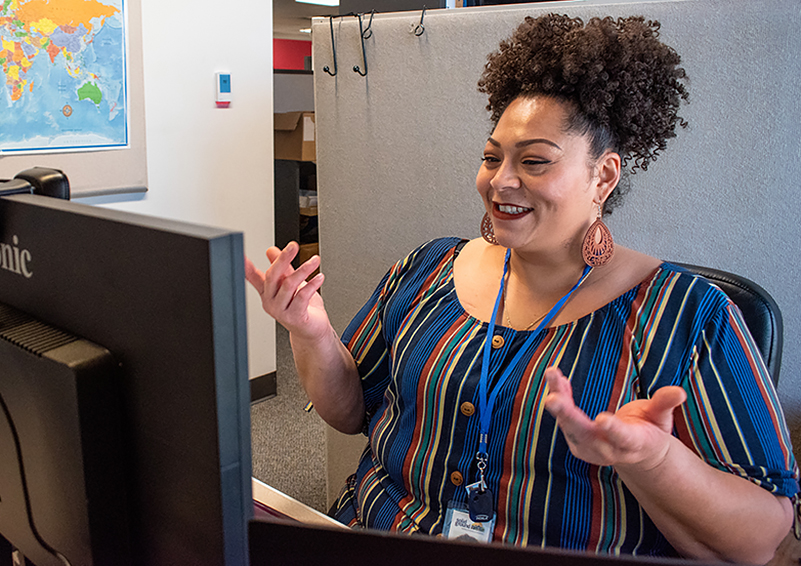 Black woman with curly hair piled on top of her head and wearing a colorful striped top sits in a cubicle in front of a computer monitor. She's smiling and gesturing with both hands.