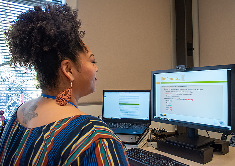 Black woman with curly hair piled on top of her head and wearing a colorful striped top sits in front of two computer screens.