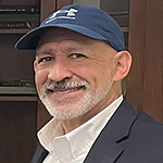 Headshot of a smiling man with white mustache and beard, wearing a blue ball cap, white-collared shirt, and black jacket.