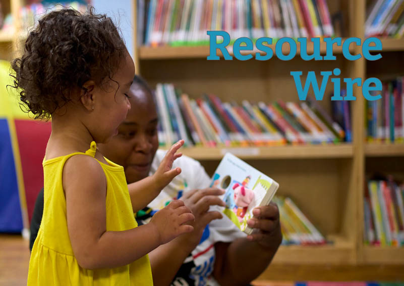 A young girl with dark, curly hair and yellow dress looks at a book with her mother in a library. The words "Resource Wire" appear above them.