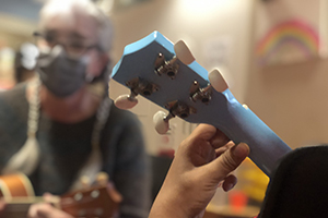 The hand of a child playing a blue ukulele is in the foreground. In the background is a woman with grey braids, glasses, and a mask demonstrates chords on the uke.