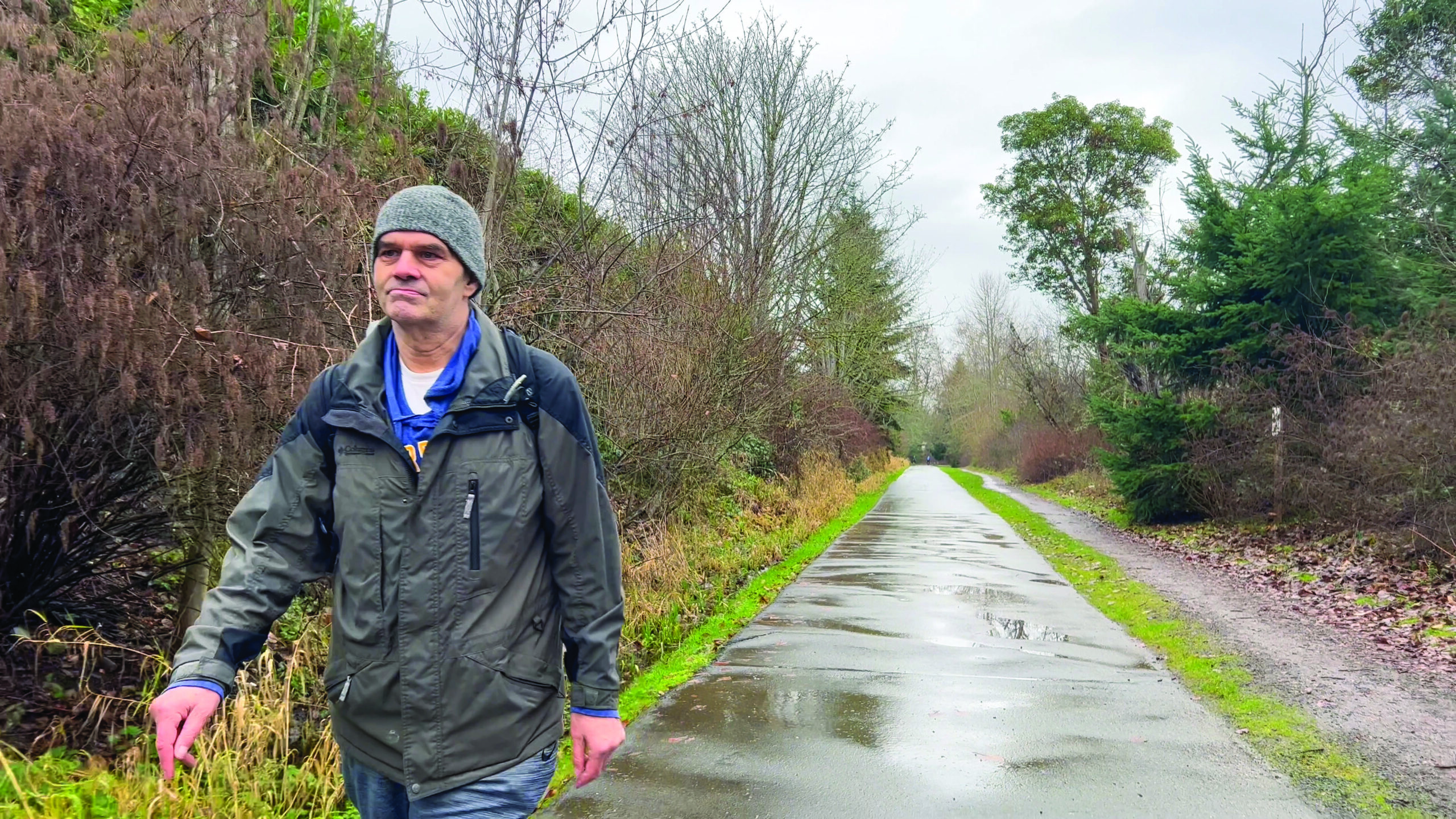 A man wearing a light grey hat, dark grey rain jacket, and jeans, strolls down a paved path wet with rain.