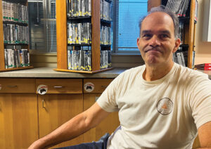 A man with greying hair, wearing an ivory T-shirt and jeans, smiles in front of several racks of DVDs.