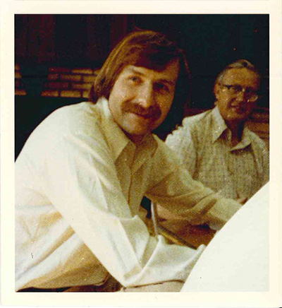 A sepia-toned photograph from the late 1980s of two men in white collared shirts sitting at a table. The man in the foreground (Frank Chopp) has long brown hair and a thick mustache.