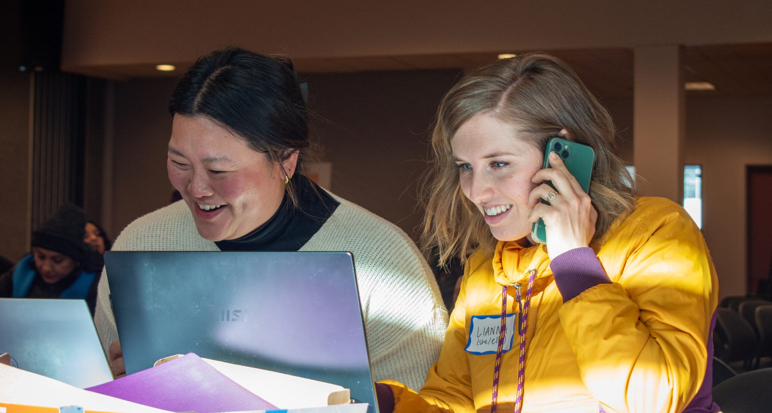 Two woman sit side by side behind laptops, one of them talking on the phone.