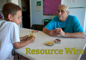 A young boy in a white tshirt plays the card game UNO with an older man in a blue tshirt. The words "Resource Wire" appear over the table in between them.