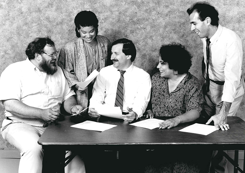 A black and white photo from the late 1980s showing five people, both men and women, in office attire sitting and standing around a table.