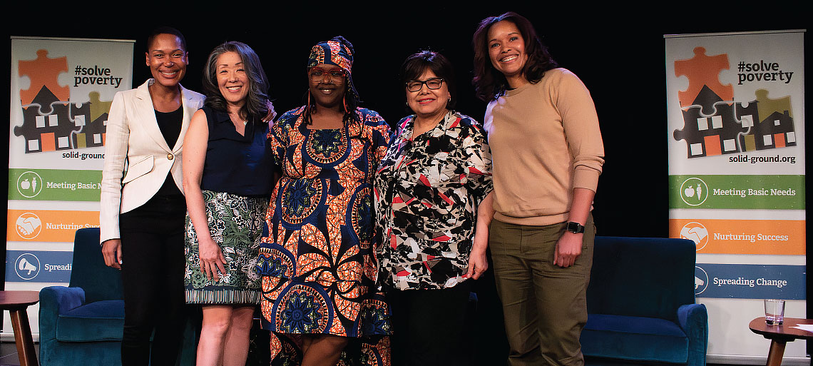 Five women of color smile from a stage with a black backdrop. On either side of them are pullup banners with #SolvePoverty above 3 puzzle pieces in Solid Ground's logo colors. Other text is solid-ground.org, Meeting Basic Needs, Nurturing Success, and Spreading Change.