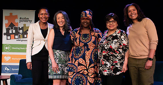 Five women of color smile from a stage with a black backdrop. On their left is a pullup banner with #SolvePoverty above 3 puzzle pieces of Solid Ground's logo. Other text is solid-ground.org, Meeting Basic Needs, Nurturing Success, and Spreading Change.