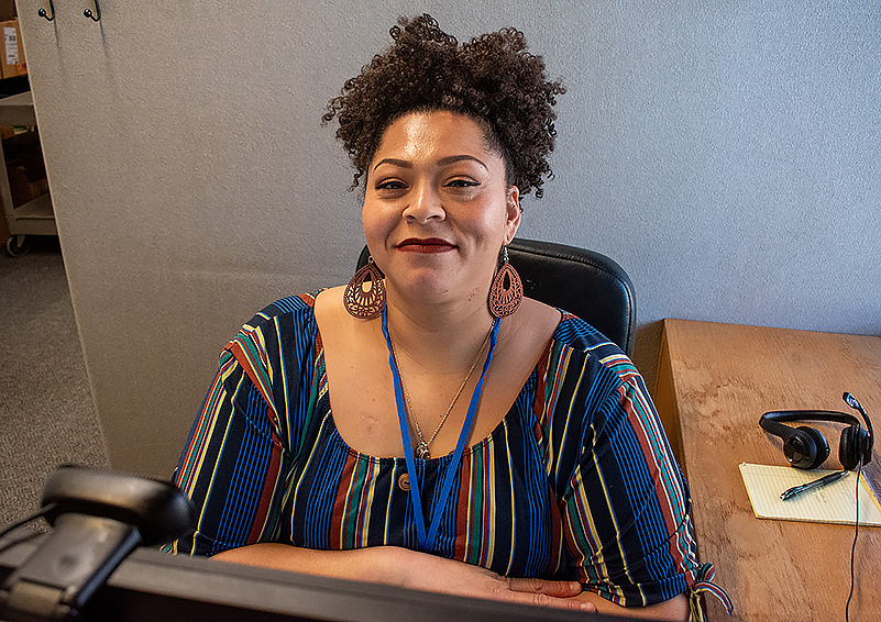 Black woman with curly hair piled on top of her head and wearing a colorful, striped top sits in a cubicle. She smiles into the camera. There's a headset, yellow note pad, and pen on the desk to her left.