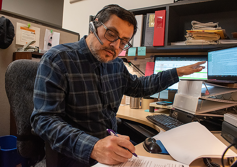 Latino man with dark glasses, hair, and facial hair – wearing a headset and a blue plaid shirt – sits at a desk in front of a computer monitor. He's pointing at the monitor with his left hand and takes notes with his right.