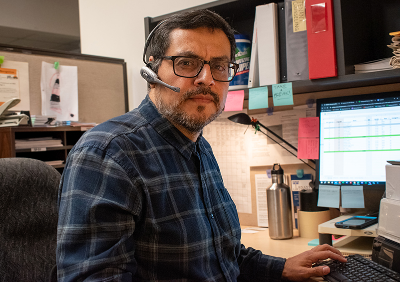 Latino man with dark glasses, hair, and facial hair – wearing a headset and a blue plaid shirt – gazes into the camera. He's sitting at a desk in front of a computer monitor and has one hand on the keyboard.