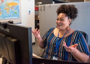 Black woman with curly hair piled on top of her head and wearing a colorful, striped top sits in a cubicle in front of a computer monitor. She's smiling and gesturing with both hands.