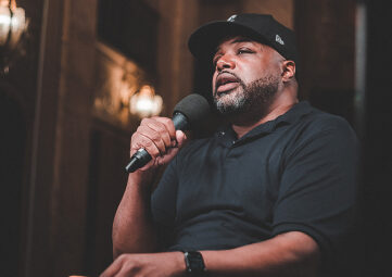 Photo of a Black man with facial hair speaking into a microphone. He wears a black cap and polo shirt.