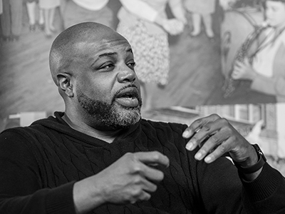 Black & white photo of a Black man with facial hair sitting in front of a backdrop depicting historical photos of Black people. He wears a black long-sleeved shirt and gestures with his hands.