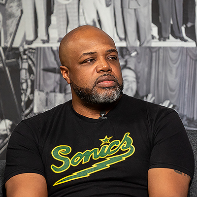 Photo of a Black man with facial hair sitting in front of a black & white backdrop depicting historical photos of Black people. He wears a black T-shirt with a green and yellow Sonics logo on it.