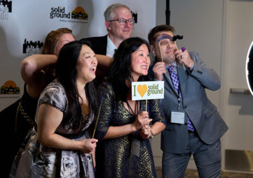 A group of people in fancy dress smile and lean into each other for a photo in front of a back drop with the Solid Ground logo on it. One person holds a sign that says "I ♥ Solid Ground."