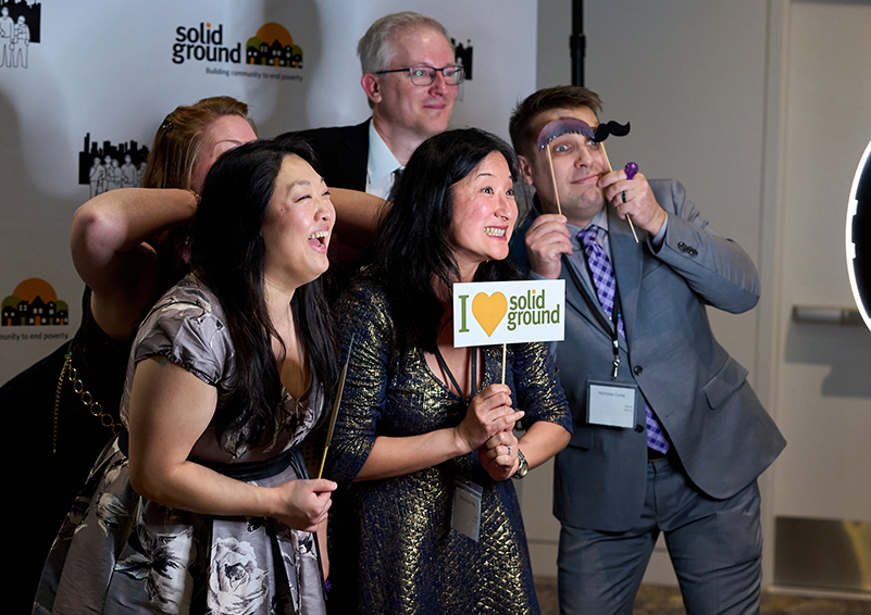 A group of people in fancy dress smile and lean into each other for a photo in front of a back drop with the Solid Ground logo on it. One person holds a sign that says "I ♥ Solid Ground."