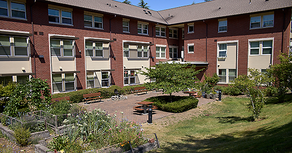 A 3-story brick apartment building, with gardens, trees, and a patio in the bend of the building.