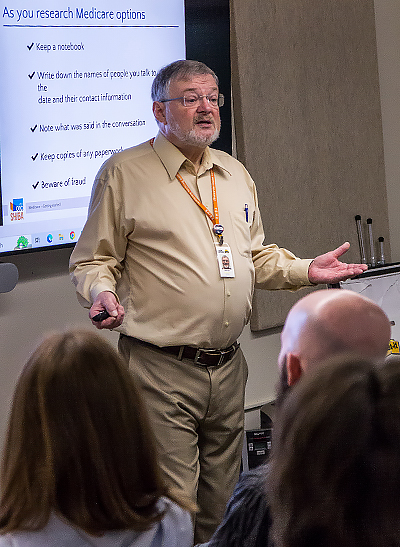 A man with grey hair and beard wearing glasses, an orange lanyard, and khaki pants and shirt, speaks to a small audience. Behind him is a PowerPoint slide titled 'As you research Medicare options.'