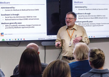 A man with grey hair and beard wearing glasses, an orange lanyard, and khaki pants and shirt, speaks to a small audience of people with grey hair. Behind him are PowerPoint slides titled 'Decide how you want to get your Medicare.'