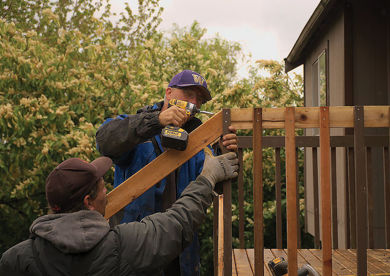 A middle-aged man wearing a purple and gold UW cap uses a drill to fix a handrail. Another man in a brown cap stand below him, holding the rail secure for him.