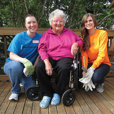 On a woodedn deck, two young women with brown hair and wearing workgloves crouch on either side of a white-haired lady wearing glasses and sitting in a wheelchair. They're all smiling broadly.