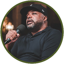 Circular photo with a forest-green frame of a Black man with facial hair speaking into a microphone. He wears a black cap and polo shirt.