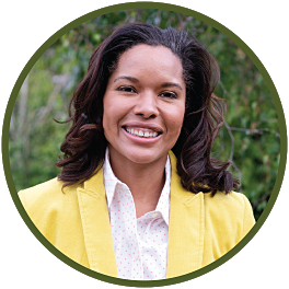 Circular photo with a forest-green frame of a Black woman with shoulder-length dark-brown hair wearing a bright yellow blazer and white blouse with small pink polka dots, standing in front of green bushes.
