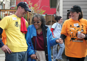 An older lady wearing sunglasses and using a cane grins at a volunteer on her left. He wears a black cap and yellow T-shirt and smiles back at her. On her right, she's holding the hand of a volunteer wearing a black Rebuilding Together cap and orange T-shirt.