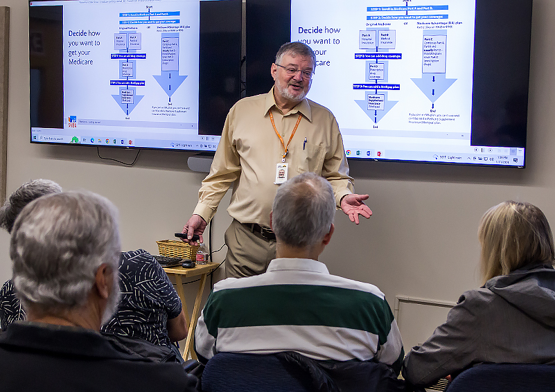 A man with grey hair and beard wearing glasses, an orange lanyard, and khaki pants and shirt, speaks to a small audience of people with grey hair. Behind him are PowerPoint slides titled 'Decide how you want to get your Medicare.'