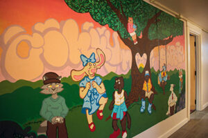 A colorful indoor mural showing cartoon animals dressed like people together under a tree with a heart-shaped hole.