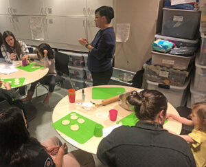 Children and adults sit around tables in a cluttered room, circles of dough on bright green cutting boards in front of them.