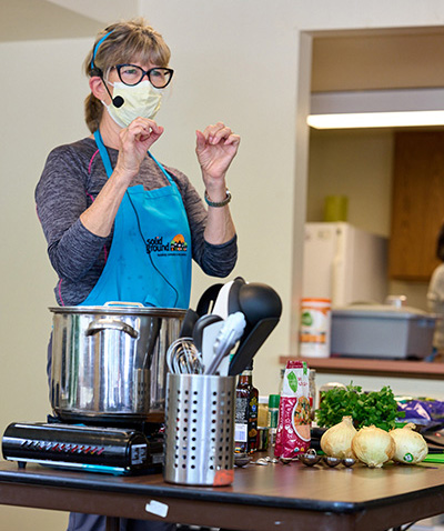 A woman wearing a teal cooking apron and a headset gestures she talks to people out of frame from behind a table with a portable stove top, cooking pot, utensils, onion, and other ingredients.