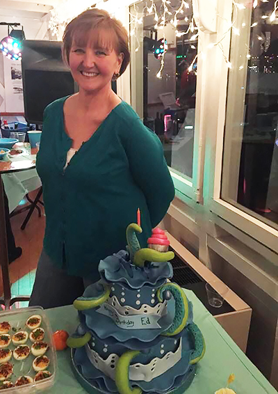 A woman smiles as she stands behind a green-and-white birthday cake shaped like a sea monster.