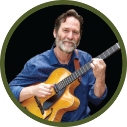 Circular photo with a forest-green frame of a white man with greying hair, nustache, and beard plays guitar. He wears a denim-blue shirt.